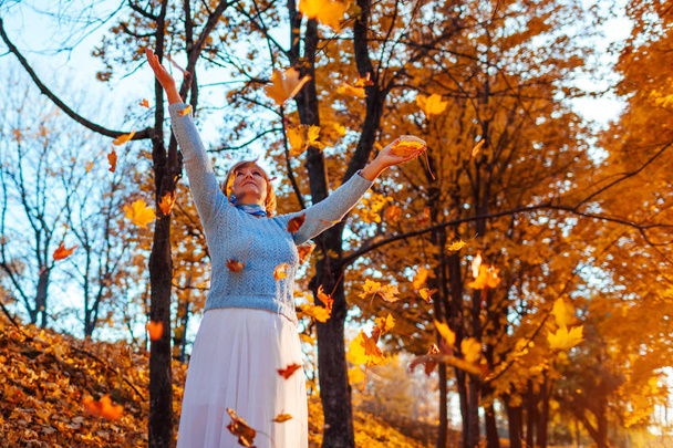 Mujer de mediana edad arrojando hojas en el bosque de otoño al atardecer. Mujer mayor divirtiéndose al aire libre
 - Foto, Imagen