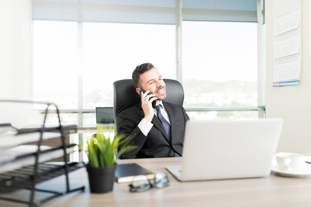 Середній дорослий підприємець у костюмі посміхається, маючи розмову зі смартфонами у власному офісі
 - Фото, зображення