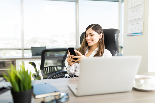 Smiling young businesswoman using mobile phone at desk in office - 写真・画像