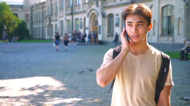 Pretty asian guy is talking over phone and looking at camera straight while standing near park, outdoor, sunny weather in city - Filmmaterial, Video