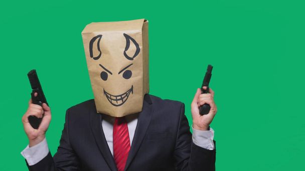 concept van emotie, gebaren. een man met een pakket op zijn hoofd, met een geschilderde smiley boos, sluwe, leedvermaak, duivel, pistolen in handen - Foto, afbeelding