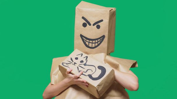 感情、ジェスチャーの概念。怒っている、スライ、塗られたスマイリーと彼の頭の上のパッケージを持つ男こきみよいと猫をなでるボックスの塗装. - 写真・画像