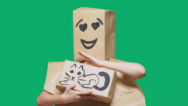 concept van emoties, gebaren. een man met een pakket op zijn hoofd, met een geschilderde emoticon, glimlach, liefdevolle ogen. speelt met een kat getrokken op de doos. - Foto, afbeelding