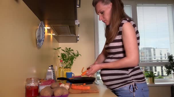 Beautiful pregnant woman peeling carrot vegetables in kitchen - Filmati, video