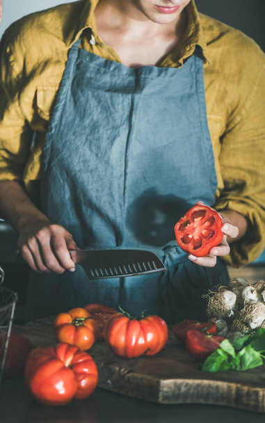 Woman in grey linen apron cooking tomato sauce, canned tomatoes or pasta with heirloom tomatoes, basil and garlic at kitchen counter. Seasonal healthy cooking, slow food or comfort food - Foto, Imagen