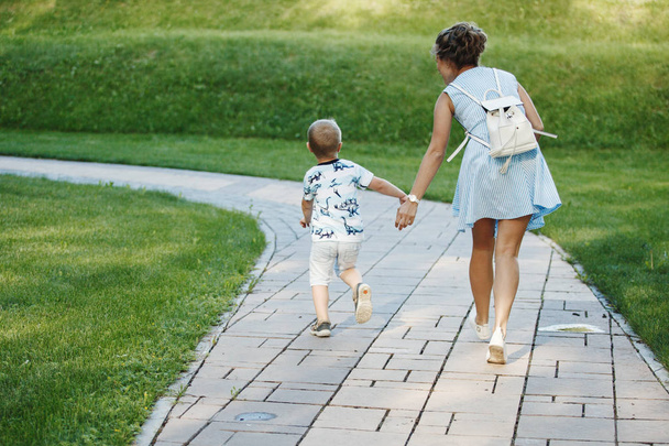 Jonge gelukkige familie: moeder in een blauwe jurk met een wit rugzak en blonde jongen lopen op een stenen bestrating op een groene weide in een park. Het concept van welzijn van de familie en een gezonde levensstijl. - Foto, afbeelding