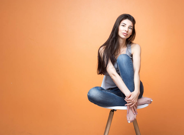 Portrait of a young girl sitting in the chair and looking into the camera against orange background.  - Foto, Bild