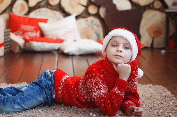 Хлопчик у різдвяних капелюхах лежить і сміється на килимі біля новорічної ялинки на фоні порізів дерев
. - Фото, зображення