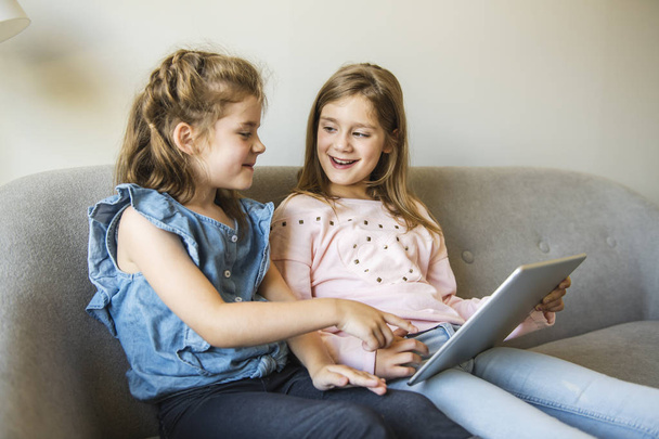 two sisters girls sit on the sofa at home with tablet - Foto, Imagem