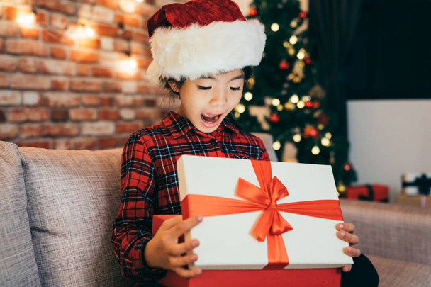 joyful cute little girl surprised open christmas gift at home. attractive girl childhood with santa hat sitting in living room. open gifts boxing day concept. - Фото, зображення