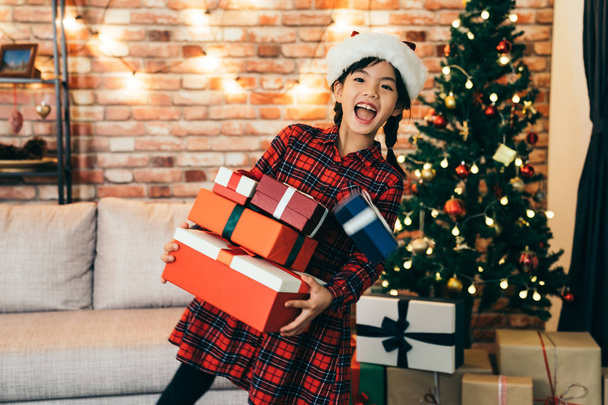 cute girl holding gift showing big toothy smile. lovely daughter with santa hat standing in front of the xmas tree. joyfully getting gifts at Christmas holiday at home. - Fotó, kép
