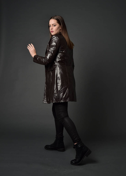 full length portrait of brunette girl wearing long leather coat and boots. standing pose with back to the camera, on grey studio background. - Foto, imagen
