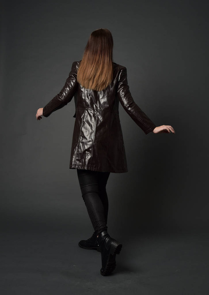 Ganzkörperporträt eines brünetten Mädchens in langem Ledermantel und Stiefeln. Stehpositionen mit dem Rücken zur Kamera, auf grauem Studiohintergrund. - Foto, Bild