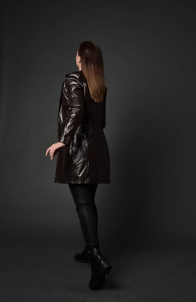 портрет дівчини брюнетки в довгому шкіряному пальто і чоботи. стояча поза ззаду до камери, на сірому студійному фоні
. - Фото, зображення