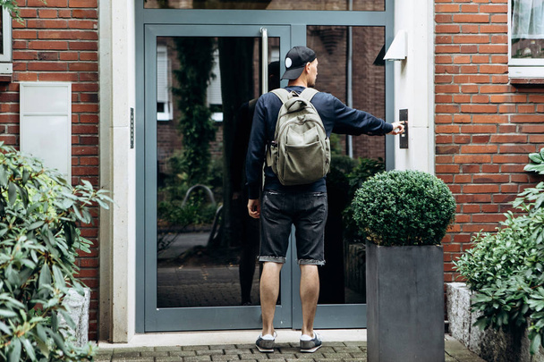 Турист звонит в дверь, чтобы зарегистрироваться в комнате он забронировал или студент с рюкзаком возвращается домой после занятий в институте или в отпуске
. - Фото, изображение