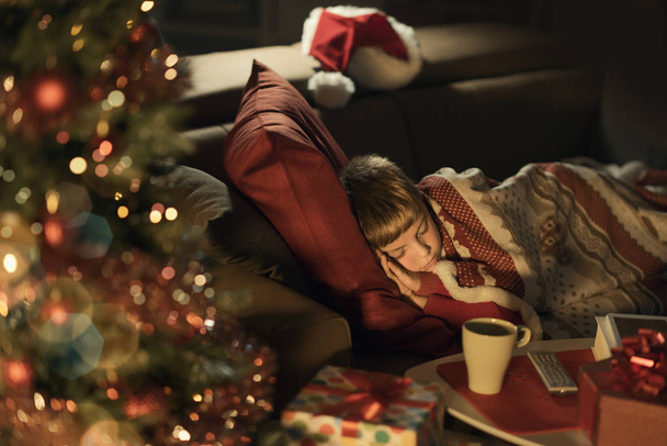 Cute little boy sleeping on the sofa next to the Christmas tree, he is waiting for Santa Claus, Christmas and holidays concept - Photo, Image