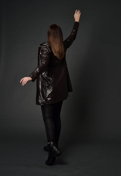 Ολόσωμο πορτρέτο μελαχρινή κοπέλα φορώντας μακρύ δερμάτινο παλτό και μπότες. θέτουν στέκεται με την πλάτη στην κάμερα, σε φόντο γκρι στούντιο. - Φωτογραφία, εικόνα