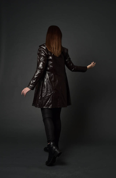 Pełna długość Portret brunetka Girl ubrany długi skórzany płaszcz i buty. Stojący poza z powrotem do aparatu, na studio szary tło. - Zdjęcie, obraz