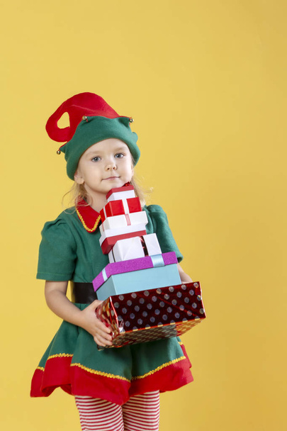 Niña en traje de elfo de Navidad con regalos sobre fondo amarillo
 - Foto, Imagen