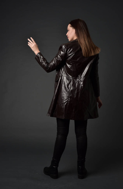 full length portrait of brunette girl wearing long leather coat and boots. standing pose with back to the camera, on grey studio background. - Foto, imagen
