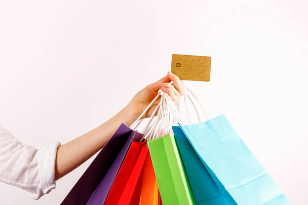 Cropped shot of female hand holding bunch of different colorful blank shopping bags with golden limitless credit card over isolated white background. Black friday sale concept. Copy space, close up. - Foto, afbeelding
