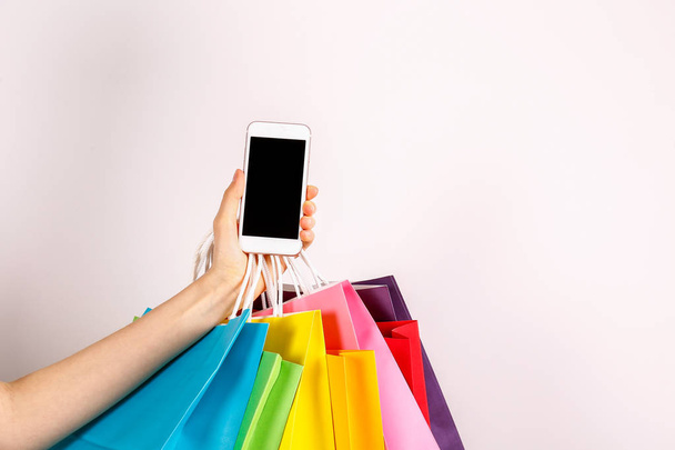 Cropped shot of female hand holding bunch of different colorful blank shopping bags and blank screen mobile phone over isolated white background. Black friday sale concept. Copy space, close up. - Foto, immagini