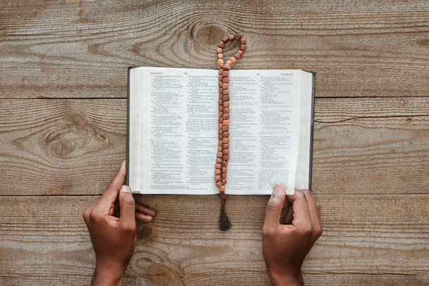 abgeschnittene Aufnahme eines afrikanisch-amerikanischen Mannes beim Lesen der Heiligen Bibel mit oben liegenden Perlen auf einem Holztisch - Foto, Bild