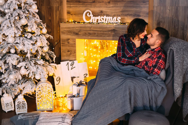 ευτυχισμένο ζευγάρι ψάχνει σε κάθε άλλη συνεδρίαση σε ένα μαλακό πολυθρόνα και καλύπτεται με μια κουβέρτα σε μια ζεστή αίθουσα κοντά το τζάκι και το χριστουγεννιάτικο δέντρο - Φωτογραφία, εικόνα