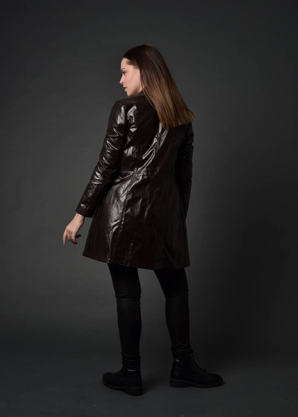 長い革のコートやブーツを身に着けているブルネットの女の子の完全な長さの肖像画。灰色のスタジオの背景に、カメラに背中を立ちポーズします。. - 写真・画像