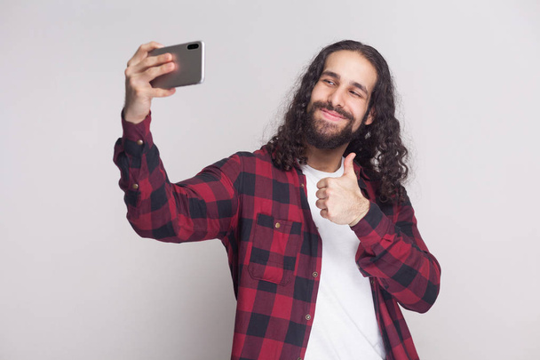 young adult man in red checkered shirt and black long curly hair standing and making selfie with smartphone and showing thumb up on grey background.  - Photo, Image