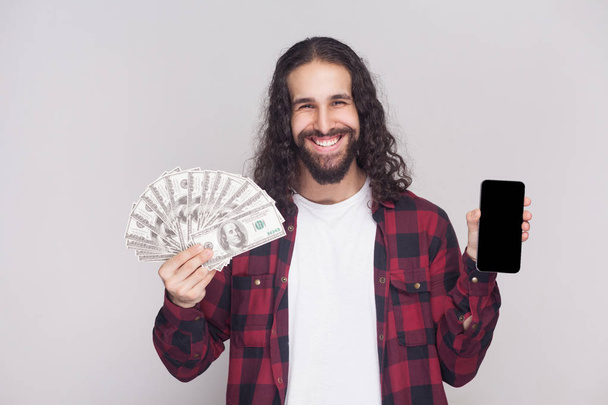 young adult happy businessman in red checkered shirt and long black curly hair showing fan of dollars and smartphone screen on grey background.  - Valokuva, kuva