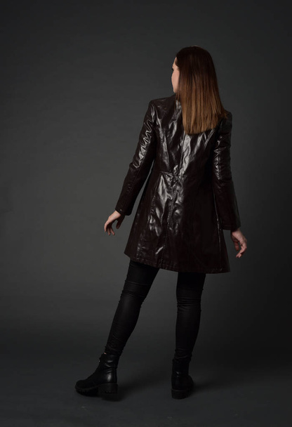 po celé délce portrét bruneta dívka, která nosí dlouhý kožený kabát a boty. postavení představují s zády ke kameře, na pozadí šedé studio. - Fotografie, Obrázek