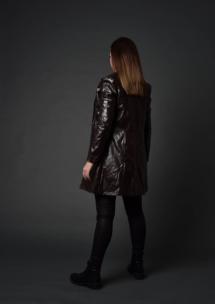 po celé délce portrét bruneta dívka, která nosí dlouhý kožený kabát a boty. postavení představují s zády ke kameře, na pozadí šedé studio. - Fotografie, Obrázek