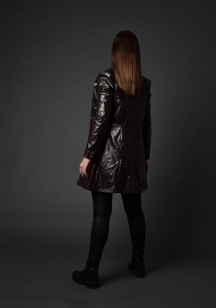 full length portrait of brunette girl wearing long leather coat and boots. standing pose with back to the camera, on grey studio background. - Photo, Image
