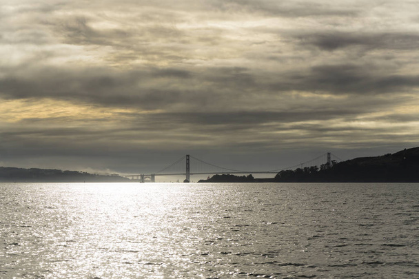 Pilvinen auringonlasku San Franciscon lahden yllä nähtynä veneestä vedessä. Näkymät kaupunkiin ja kultainen portti silta
 - Valokuva, kuva