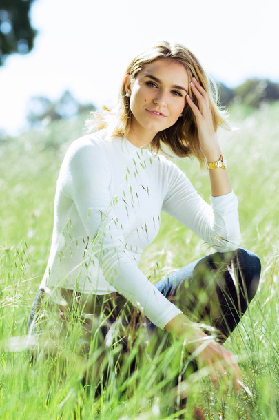 Atractiva joven rubia caucásica mujer en top blanco apretado y denim posando fuera en hierba larga
 - Foto, Imagen