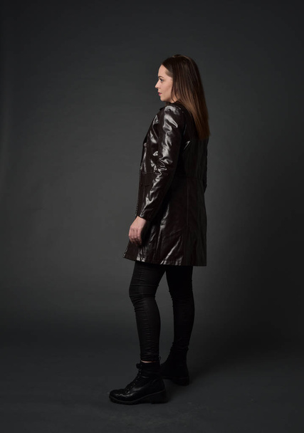 full length portrait of brunette girl wearing long leather coat and boots. standing pose with back to the camera, on grey studio background. - Photo, Image