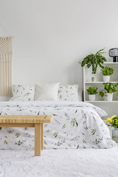 White bedroom interior with fresh plants on rack, floral sheets on double bed, carpet on the floor and empty place for your poster on the wall - Foto, afbeelding