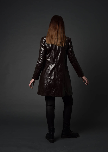 full length portrait of brunette girl wearing long leather coat and boots. standing pose with back to the camera, on grey studio background. - Foto, Imagen