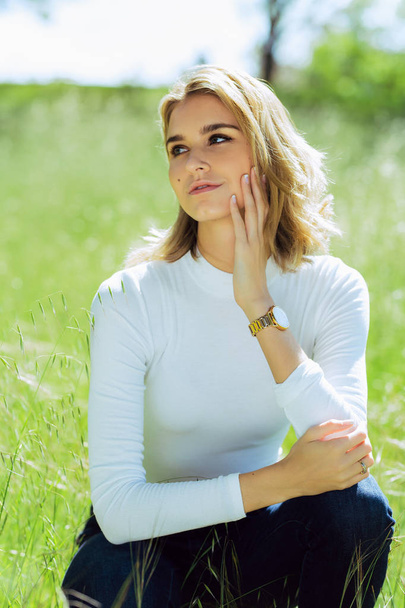 Attractive young blonde Caucasian woman in tight white top and denim posing outside in long grass - Photo, Image
