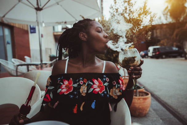 Portrait of a charming African girl sitting in a street cafe and making smoke in the glass of cocktail; young black female outdoors in a bar play with a hookah while drinking delicious mojito - Foto, Imagem