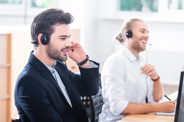 lachende mannelijke call center exploitanten in hoofdtelefoons op werkplek op kantoor - Foto, afbeelding