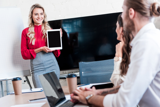 usměvavá podnikatelka zobrazeno tablet s prázdnou obrazovkou kolegům během setkání v kanceláři - Fotografie, Obrázek