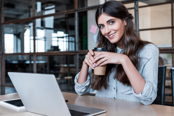 happy young businesswoman holding paper cup and smiling at camera while working with laptop in office - Fotó, kép