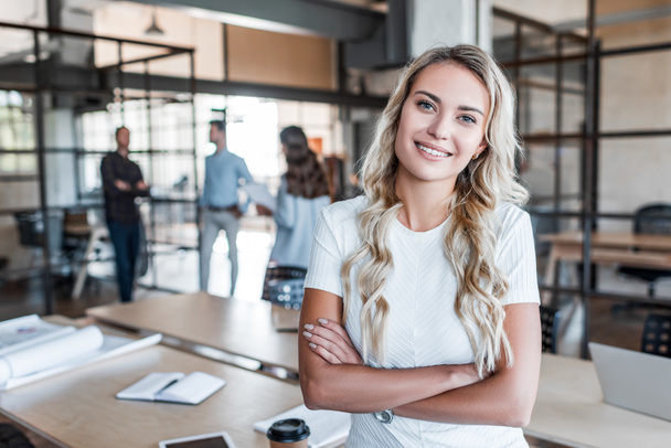 красивая молодая деловая женщина, стоящая со скрещенными руками и улыбающаяся в офисе перед камерой
 - Фото, изображение