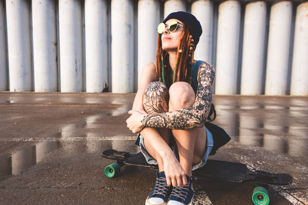 young girl with tattoos and dreadlocks in a blue cap sits on a longboard against the background of concrete structure - Фото, зображення