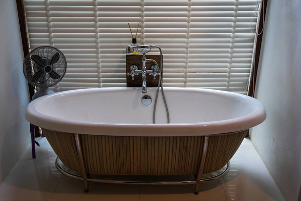 Empty white ceramic bathtub with wooden chick blind background and old electric fan. - Photo, Image