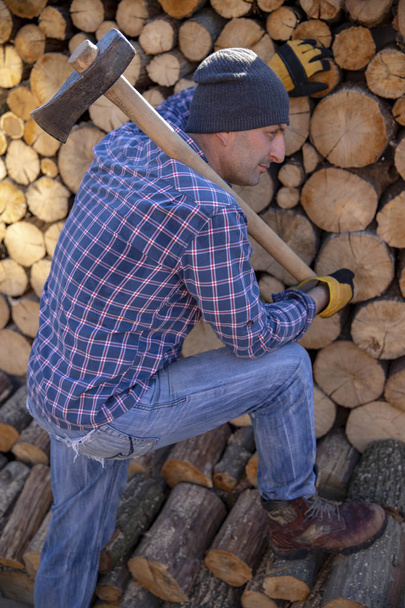 Man holding an industrial ax. Ax in hand. A strong man holds an ax in his hands against the background of chainsaws and firewood. Strong man lumberjack with an ax in his hand. Chainsaw close up. - Фото, изображение