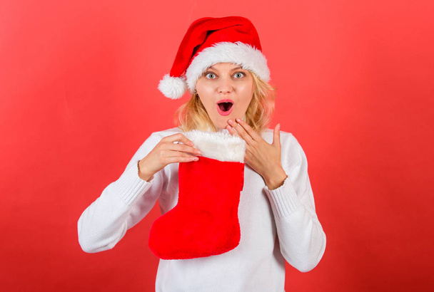 Girl cheerful face got gift in christmas sock. Traditional winter holiday. Christmas stocking concept. Check contents of christmas stocking. Woman in santa hat hold christmas gift red background - Foto, immagini
