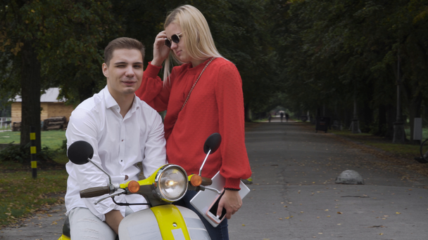 Handsome guy on a scooter is talking to a girl. Young guy in the white shirt on a yellow retro motor bicycle. Beautiful girl stands talking to a guy on a moped. Young girl with blond hair and a red - Footage, Video